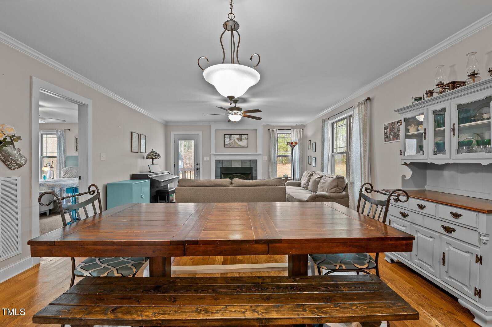 Dining area with a wooden table and two chairs in the foreground, adjacent to a living room with a sofa, coffee table, fireplace, piano, and blue hutch, all with hardwood floors and neutral-colored walls.