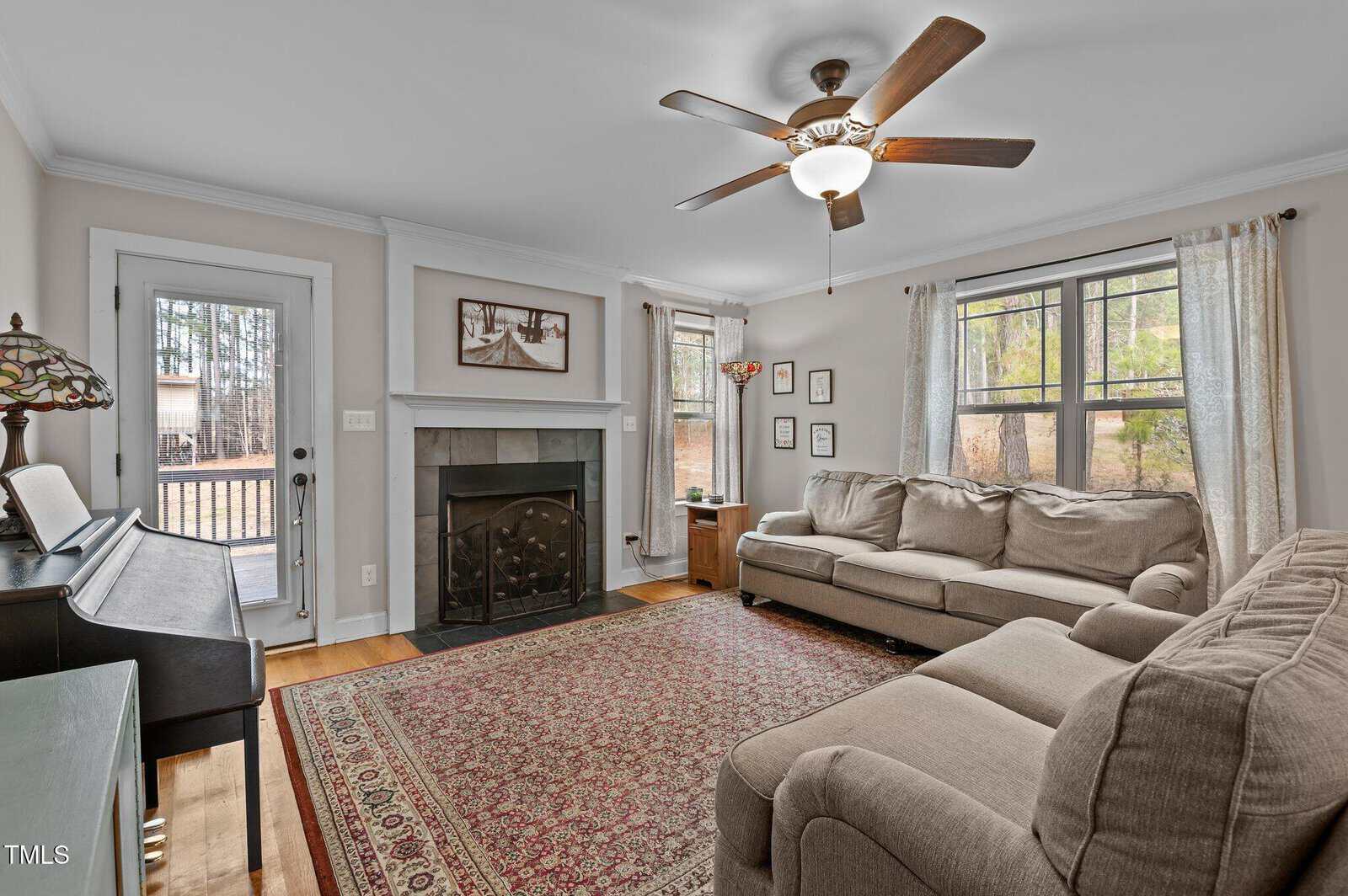 A living room with a large rug, sectional sofa, fireplace with a screen, upright piano, ceiling fan, and two windows with curtains. Framed pictures hang on the wall and a glass door opens to an outdoor area.