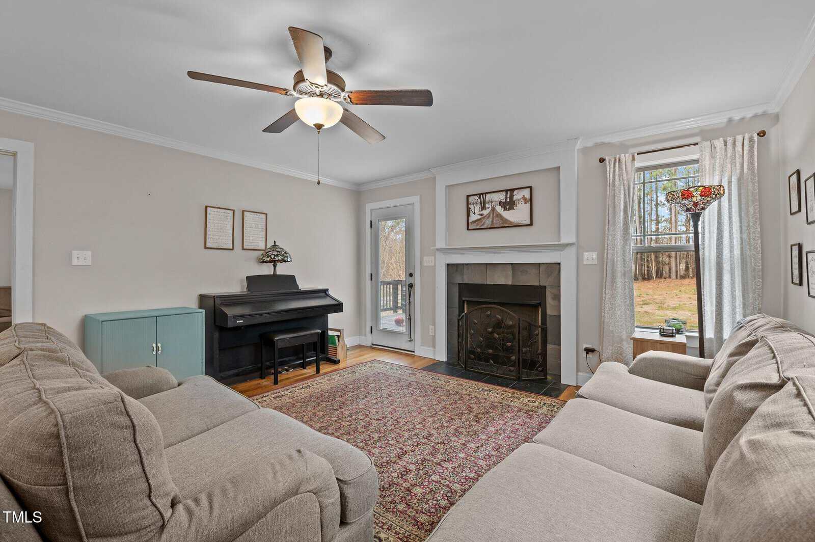 A living room with two beige sofas, a patterned rug, a fireplace with a black screen, a black upright piano, ceiling fan, window, glass door, and light-colored curtains with a backyard view.