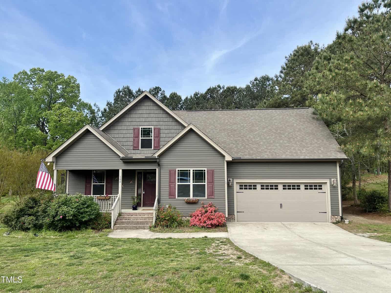 A two-story, gray suburban house with a front porch, burgundy shutters, a double garage, a concrete driveway, an American flag, and bushes and small flowers in the front yard under a blue sky.