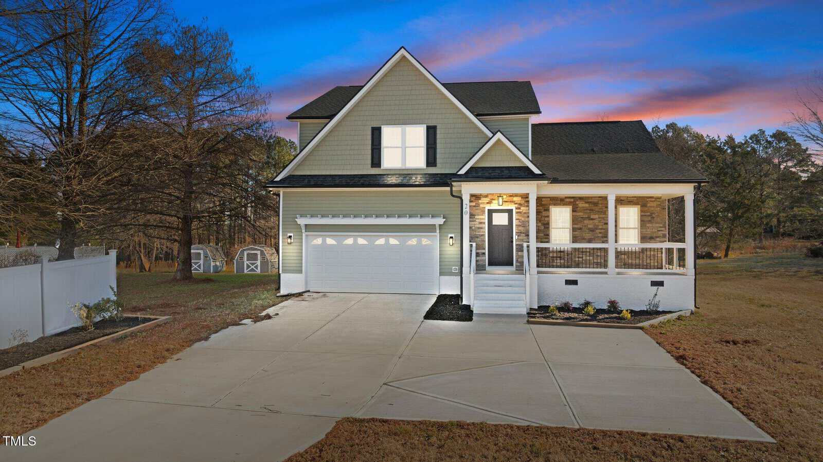 A two-story house with a double garage, front porch, and gabled roof is shown at dusk. The driveway is wide, and the yard is mostly bare. There are trees and two sheds in the background.