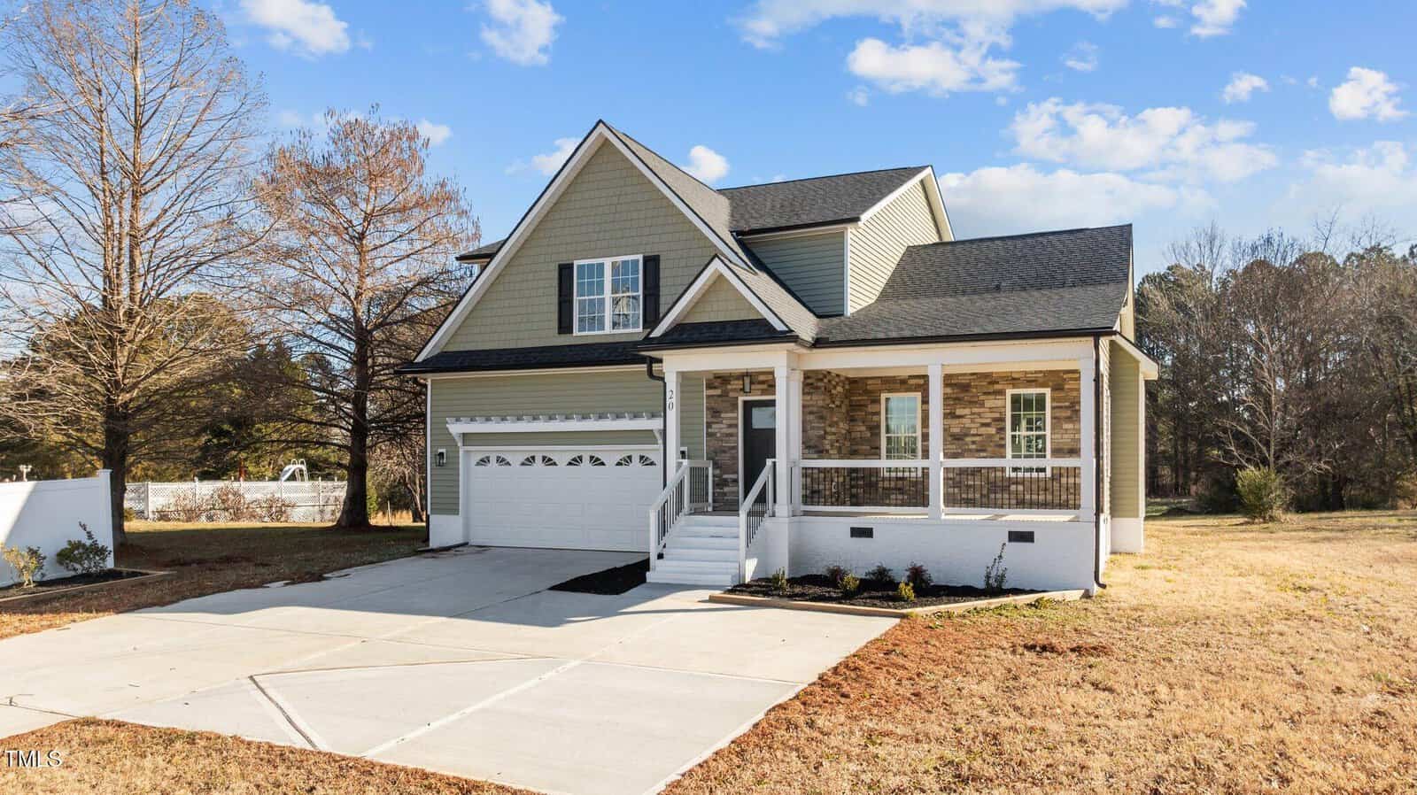 A two-story house with green siding, stone accents, a covered front porch, white trim, and an attached two-car garage. The house is surrounded by dry grass and trees under a partly cloudy sky.