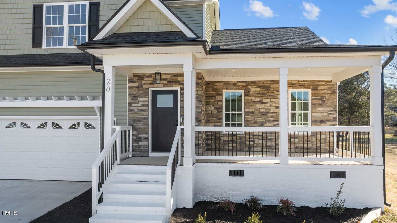 A modern house with green siding, stone accent walls, a covered front porch with white railings, a black front door, and a two-car white garage. Steps lead up to the porch. Blue sky with scattered clouds in the background.