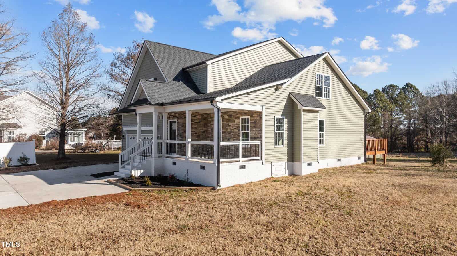 A beige two-story house with white trim and a screened-in porch stands on a lawn with dry grass. The house has a dark roof, driveway, and stairs leading to the porch. Trees and blue sky with clouds are in the background.