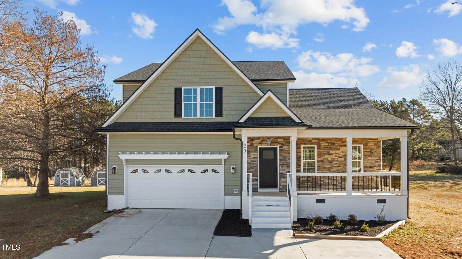A two-story suburban house with green siding, stone accents, a front porch, white garage door, and a concrete driveway. The yard has sparse landscaping and two sheds are visible in the background.
