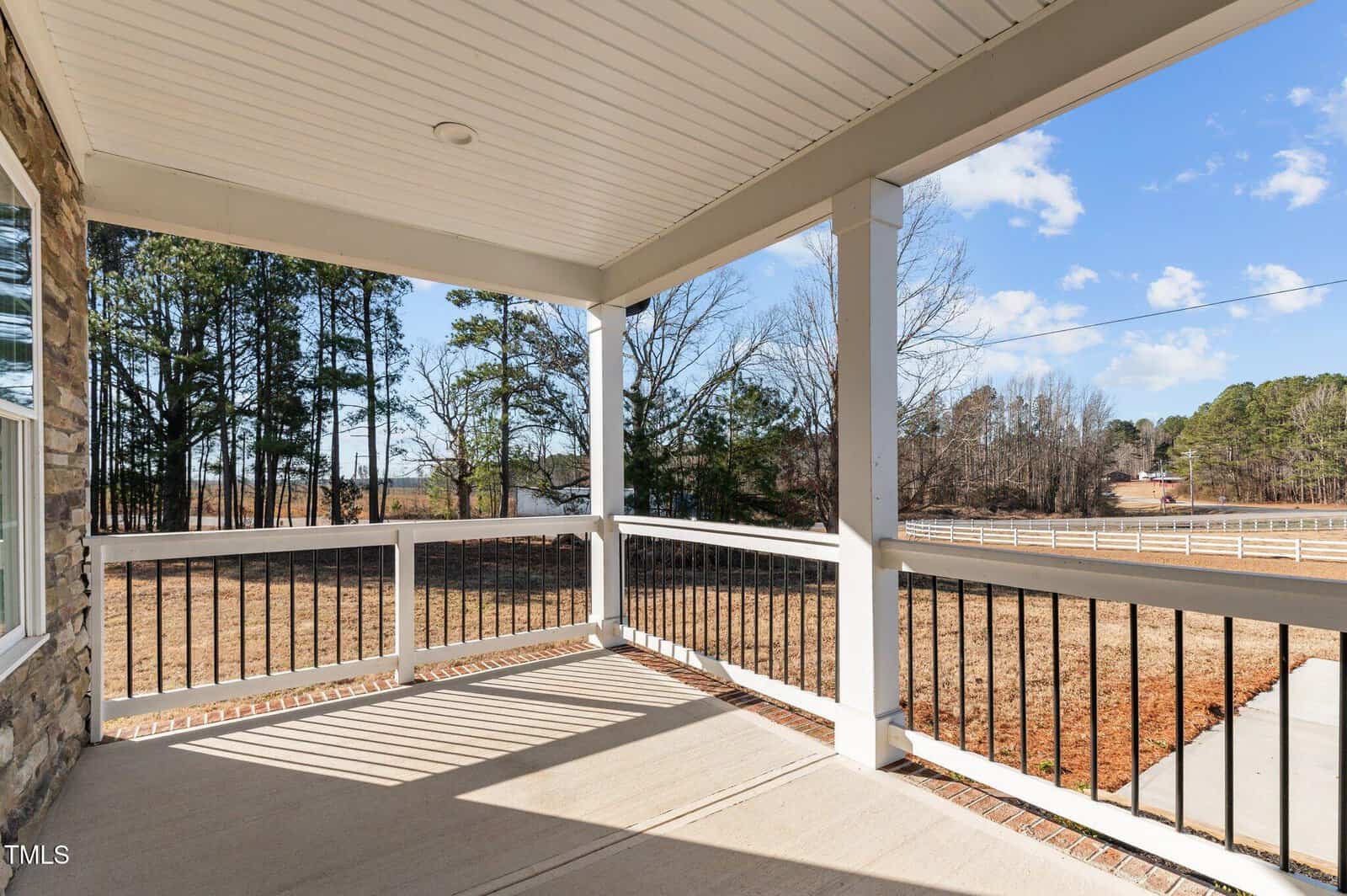 A covered front porch with white railings and black balusters overlooks a dry lawn, a road, and trees under a partly cloudy blue sky. Sunlight casts shadows on the concrete floor.
