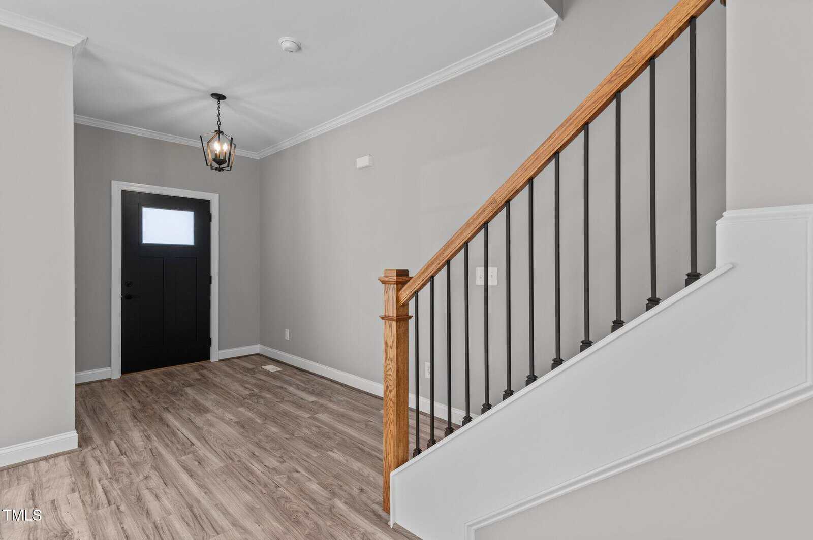 A hallway with light wood flooring, gray walls, a black front door with a frosted window, and a staircase featuring a wooden handrail and black balusters. A modern pendant light hangs from the ceiling.