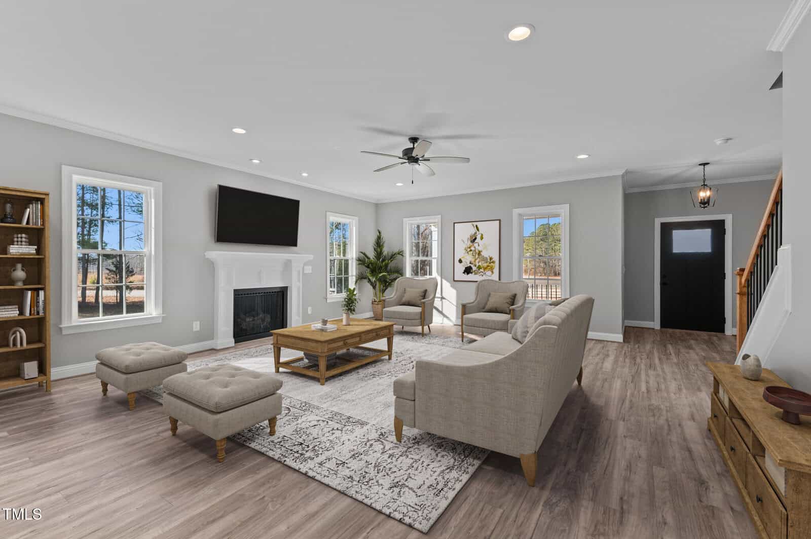 Spacious living room with light gray walls, wood flooring, large windows, a fireplace with TV above, beige sofas, a wooden coffee table, and two ottomans. Stairs and a black door are visible on the right.