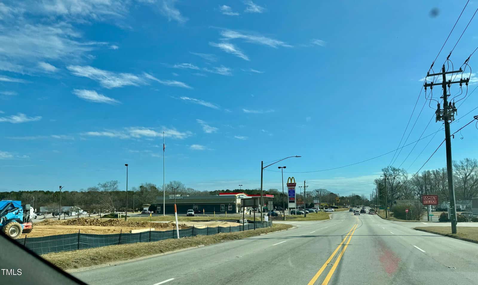 View of an empty road with a construction site on the left, featuring a blue excavator. In the background, there is a restaurant and fast-food establishment with signage. The sky is clear with scattered clouds.