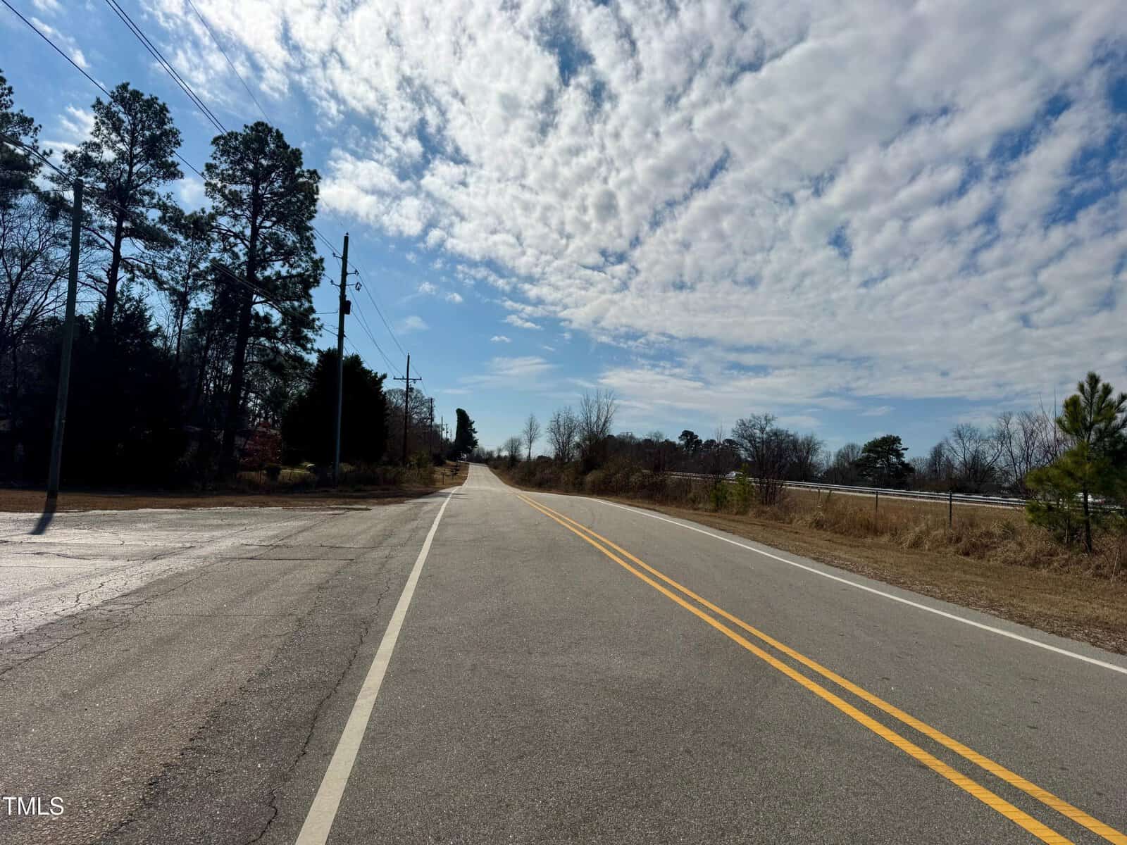 A rural two-lane road with yellow lines runs straight ahead under a partly cloudy sky. Trees line the road on both sides, and power lines are visible. The scene is peaceful and open, with light traffic and natural surroundings.