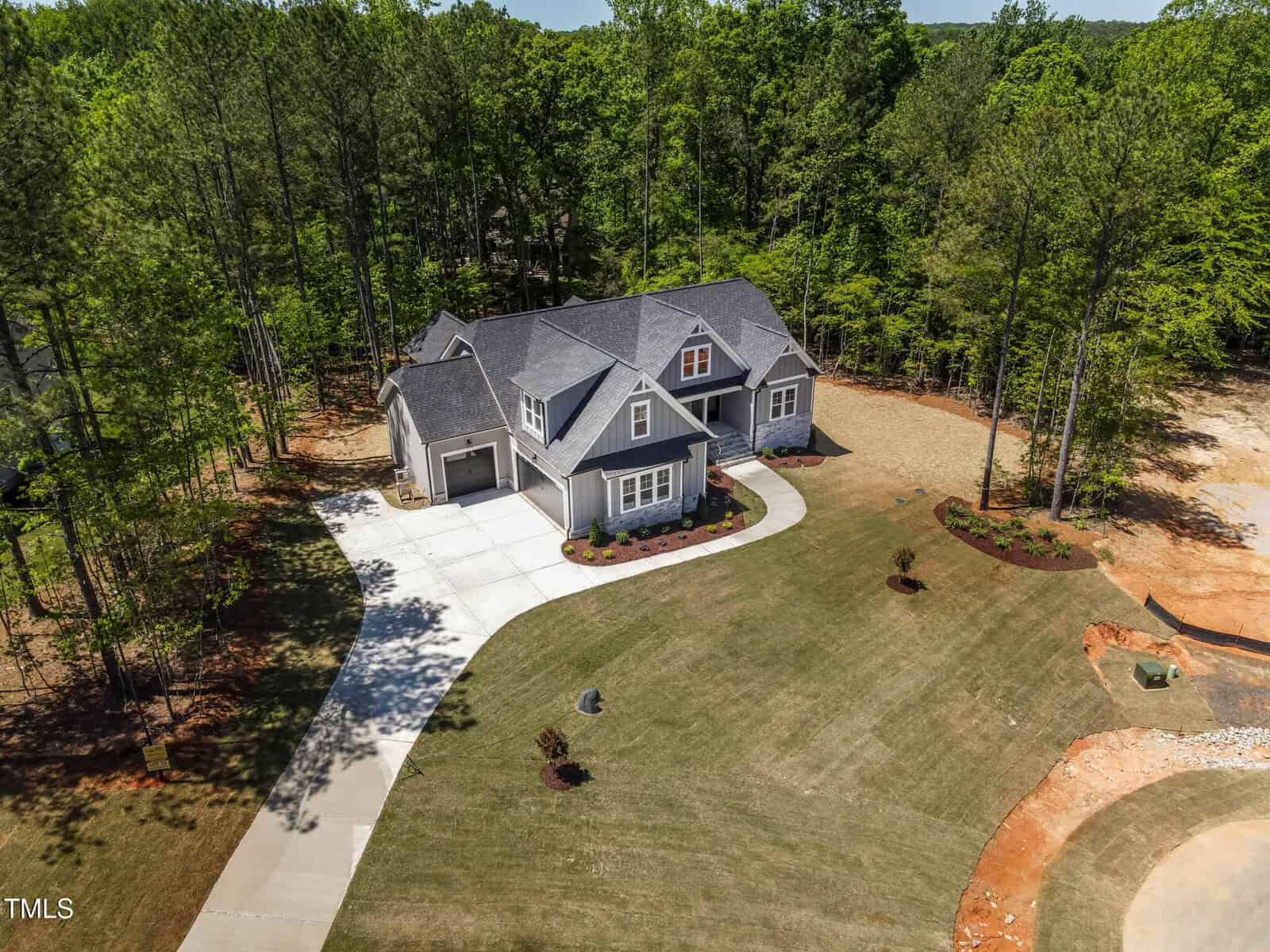 A two-story house with a gray roof and attached garage sits on a large, neatly landscaped yard surrounded by tall trees. The driveway curves to the street, and there is a patch of bare ground near the lawn.