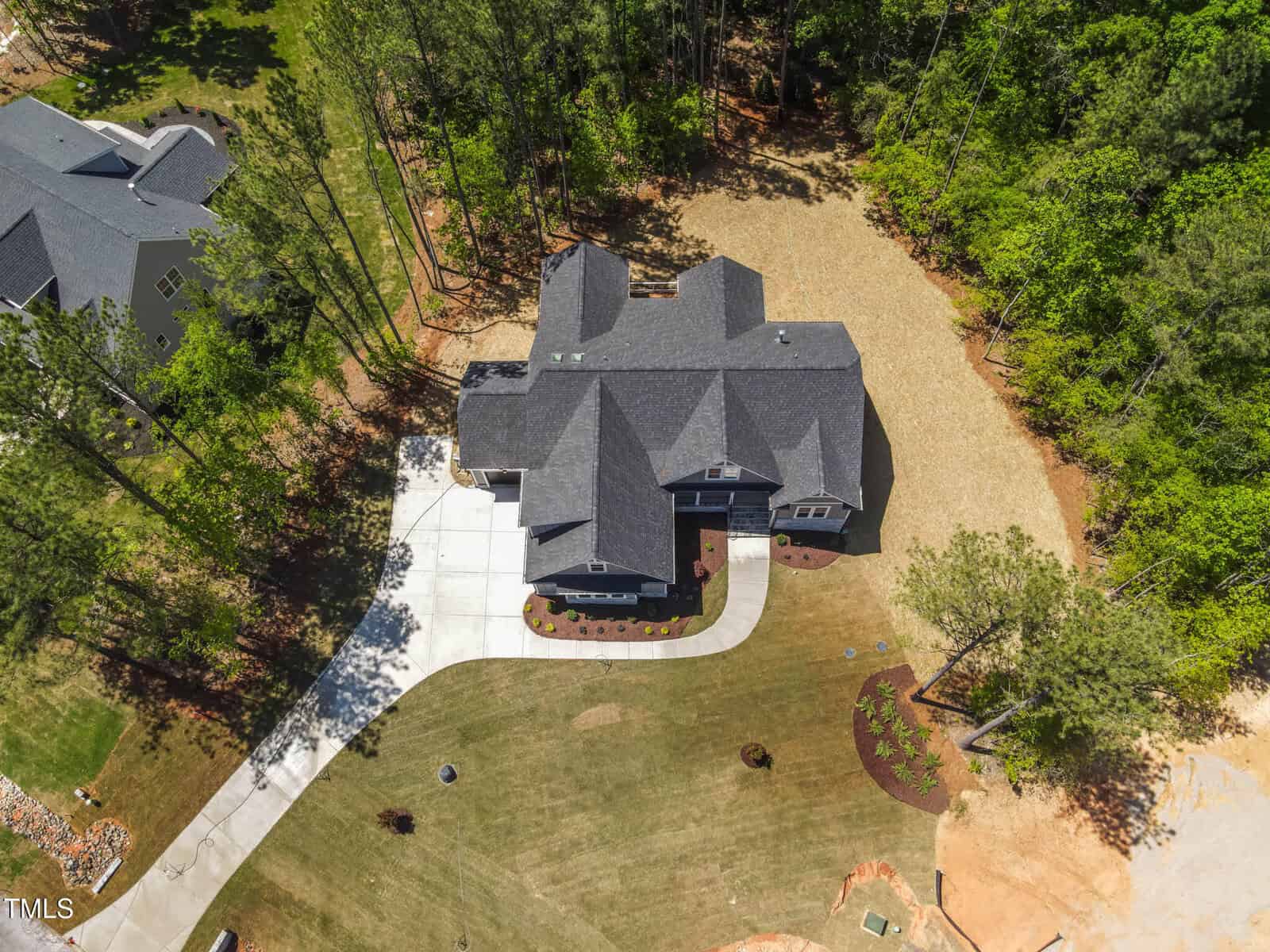 Aerial view of a large two-story house with dark roof, surrounded by grass lawn and trees. The driveway curves from the street to the attached garage. The property is bordered by wooded areas on two sides.