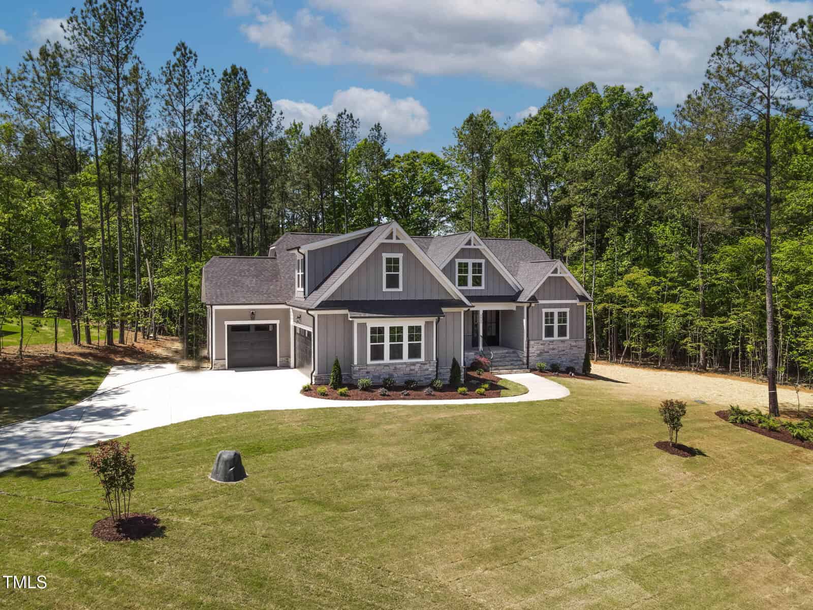 A modern gray house with a front porch and attached garage sits on a large, well-kept lawn surrounded by tall trees. A curved driveway leads to the house, and small shrubs are planted near the entrance.