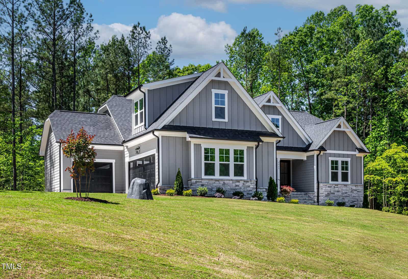 A modern gray house with white trim sits on a sloped lawn. The home has multiple gables, stone accents, a front porch, and is surrounded by trees and landscaped shrubs.