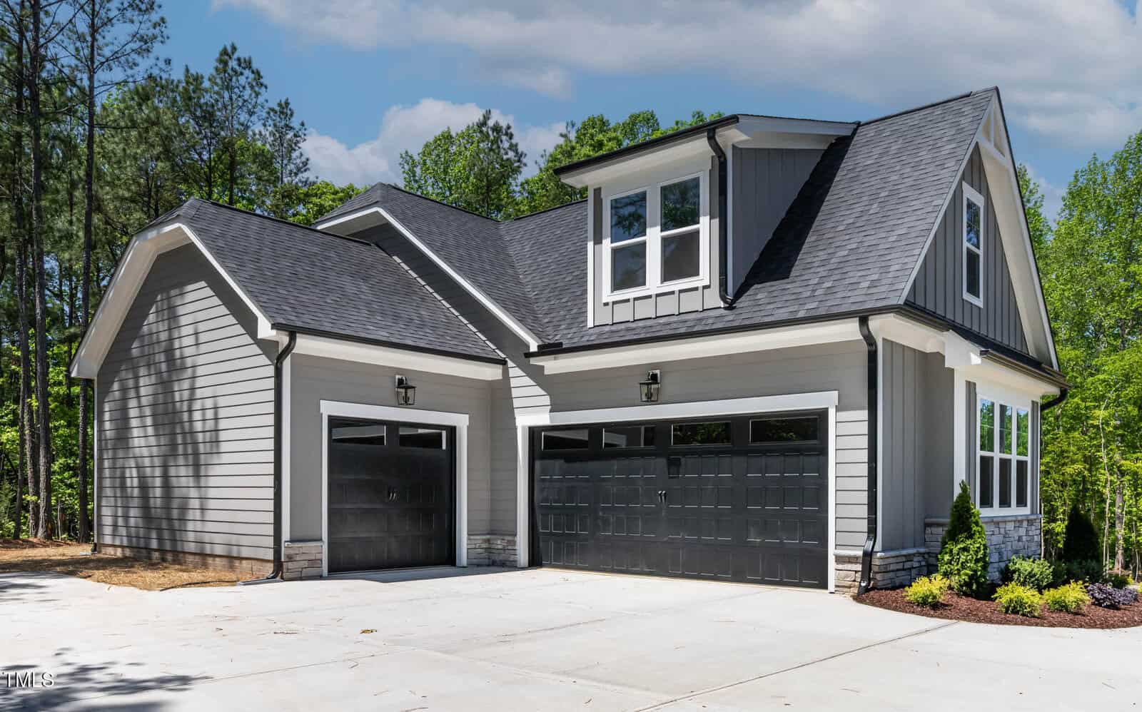 A modern two-story house with gray siding, black framed garage doors, and a large driveway, surrounded by trees and bushes under a partly cloudy sky.