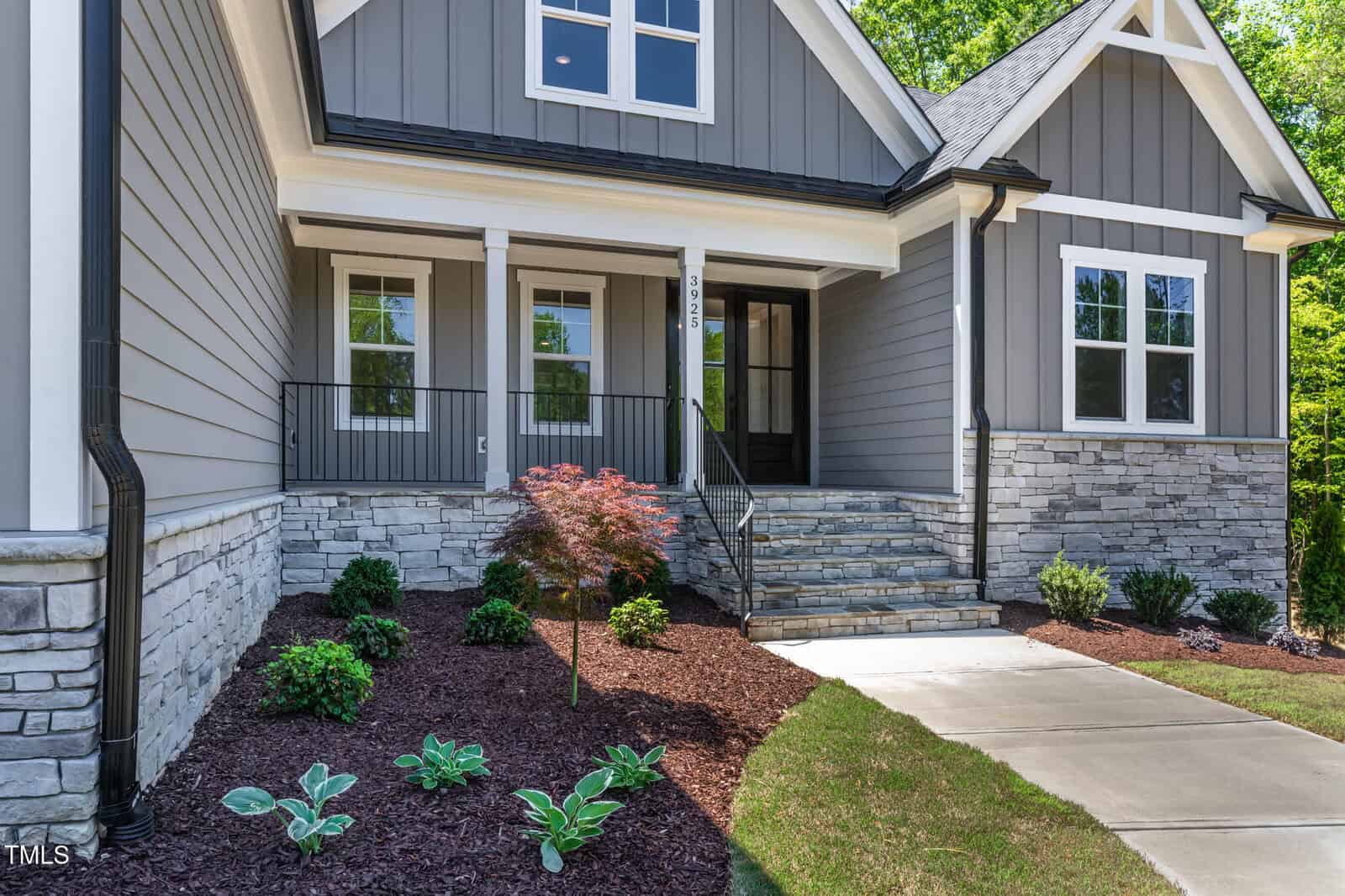 A modern house exterior with gray siding, white trim, stone accents, a covered front porch with railing, a small garden with shrubs and a young tree, and a concrete walkway leading to the steps.