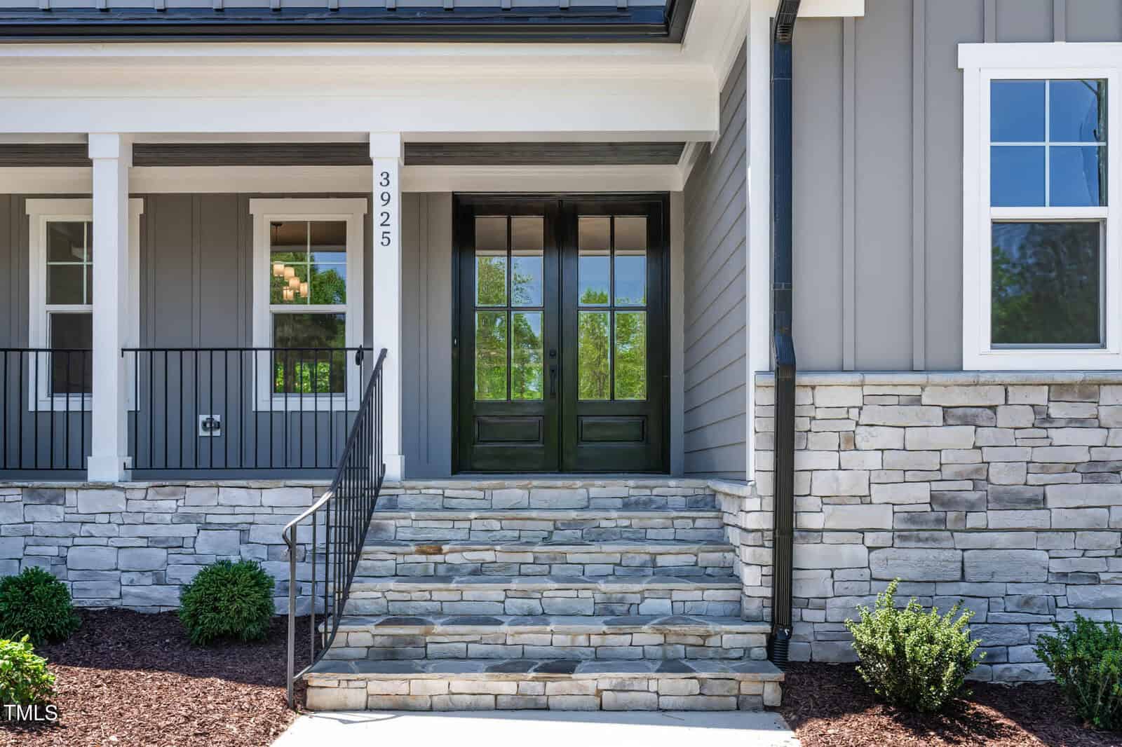 Front entrance of a house with stone steps leading to double black doors, gray siding, white trim, a small porch with metal railing, and landscaped shrubs along the stone foundation. House number 3925 is visible near the door.
