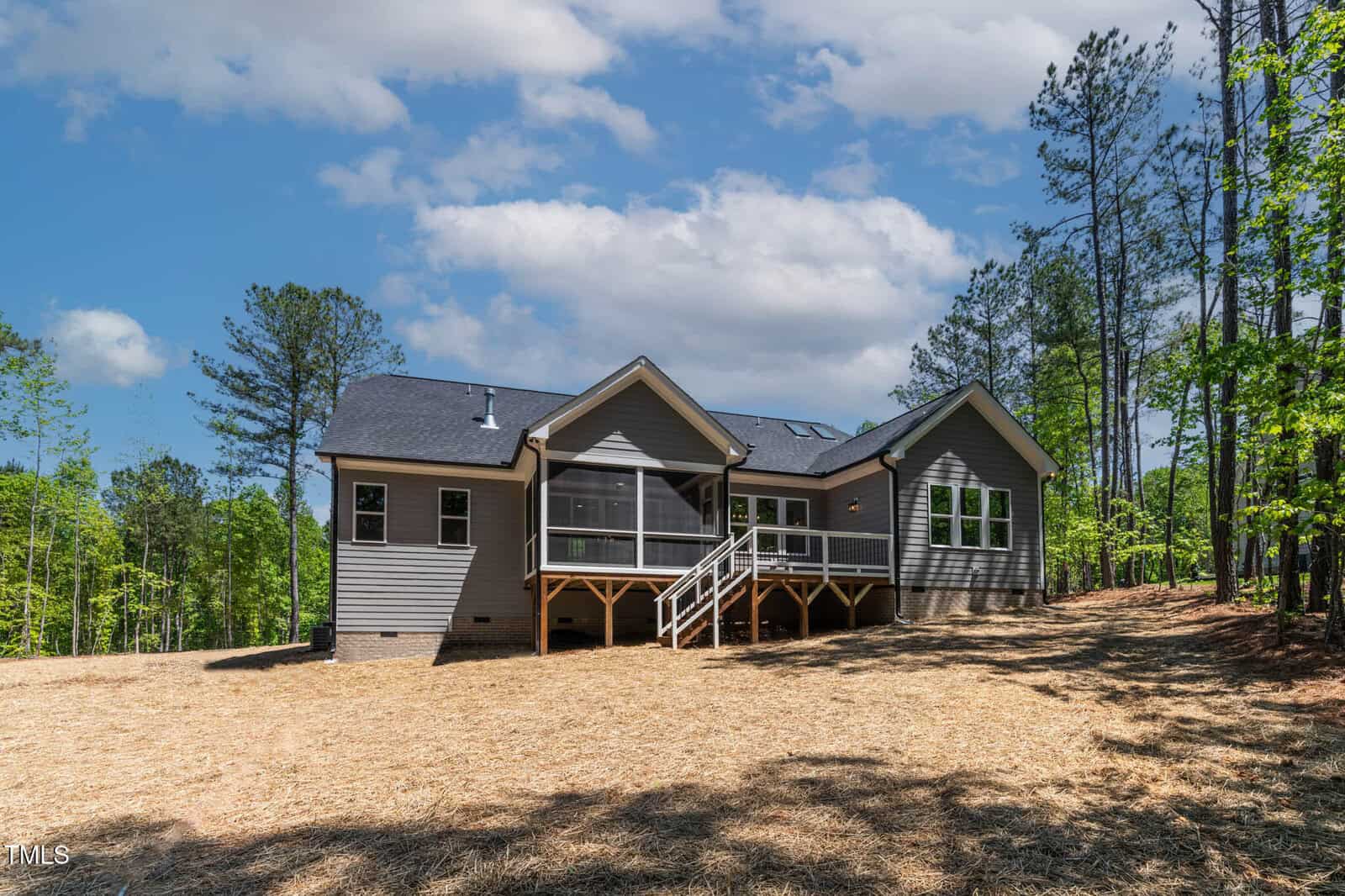 A single-story house with gray siding and a dark roof stands on a cleared lot with straw-covered ground. There is a screened-in porch with steps leading down to the yard, and trees surround the property.