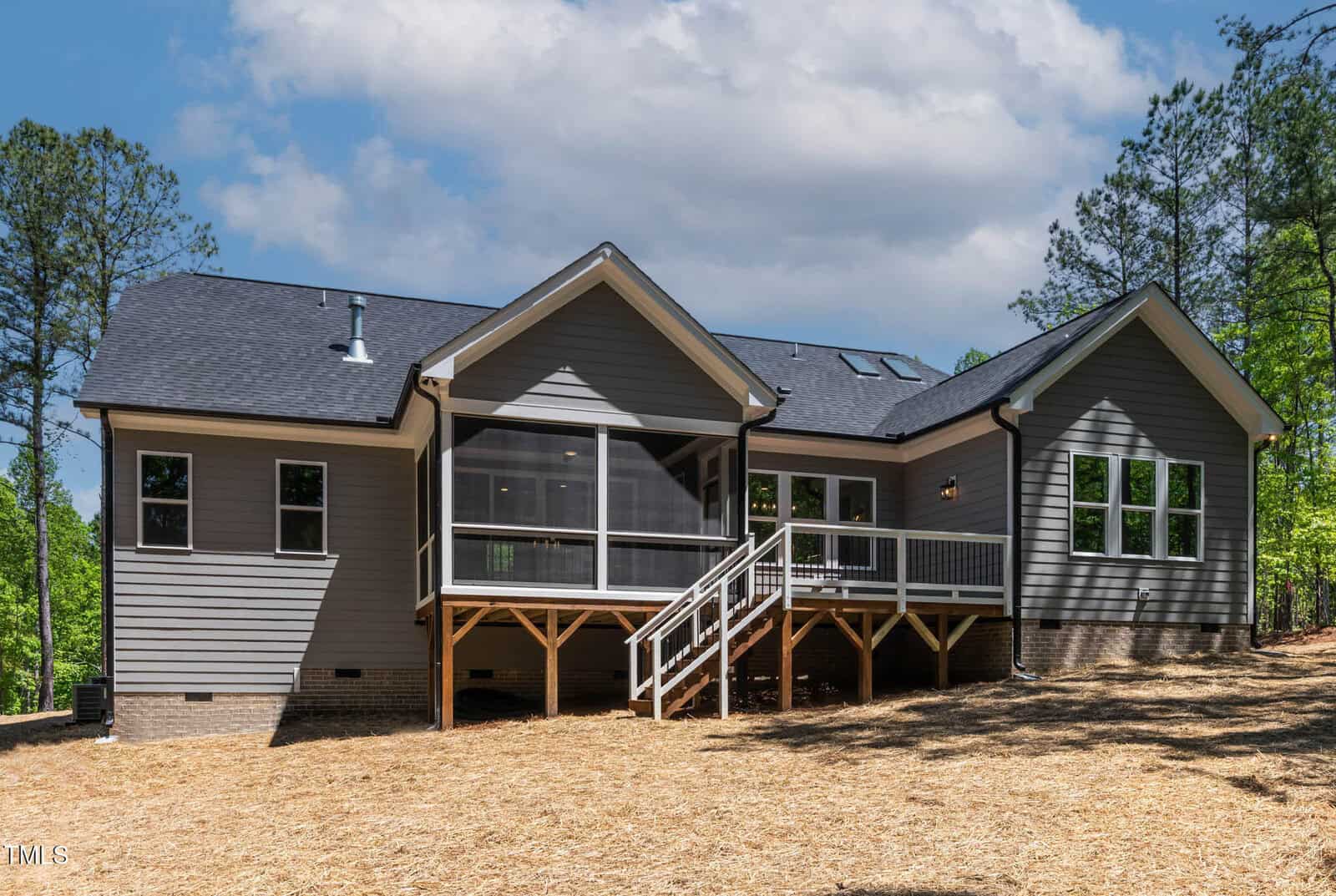 A modern, single-story house with gray siding, multiple gables, a screened-in porch, deck with stairs, and large windows. The yard is covered with straw mulch and surrounded by trees under a partly cloudy sky.