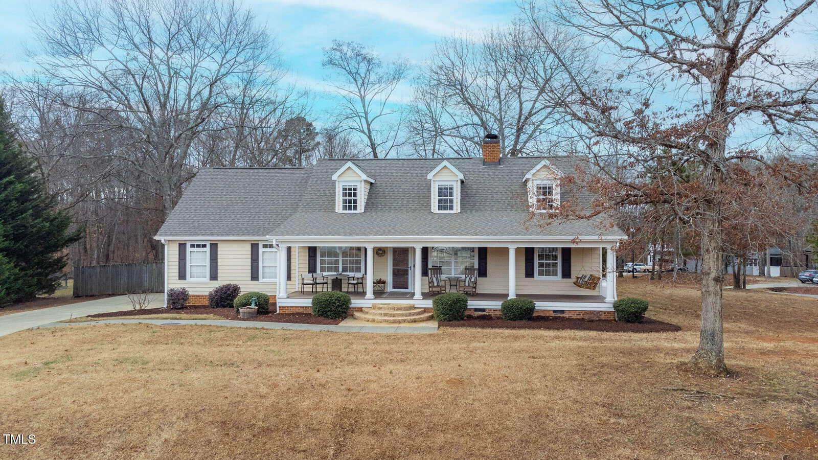 A beige, one-and-a-half-story suburban house with dormer windows, a covered front porch, and a shingled roof. The yard is mostly brown grass with sparse shrubs and trees, and a driveway runs along the left side.