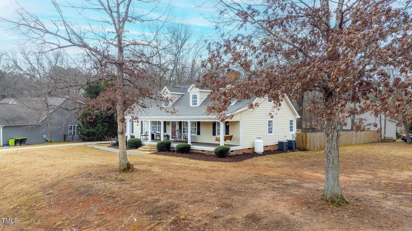 A light yellow house with white trim and a covered front porch sits on a large, mostly bare yard with a few trees. The house has multiple gables and dormer windows, with a wooden fence on one side and garages nearby.