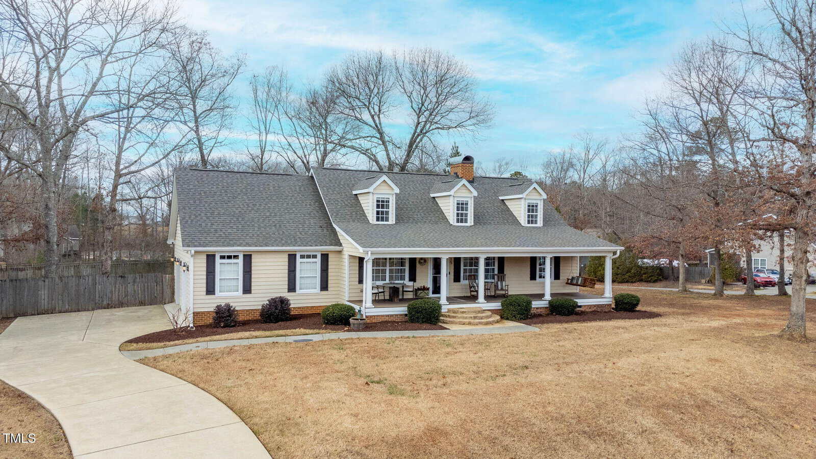 A beige single-story house with dormer windows and a covered front porch sits on a large lot with a curved driveway, manicured bushes, and trees in the background under a partly cloudy sky.