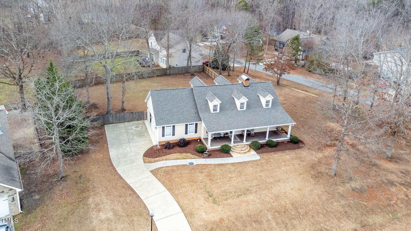 Aerial view of a single-story house with a covered front porch, dormer windows, and a curved driveway on a large lot with sparse grass and surrounding trees. Other houses and a road are visible in the background.