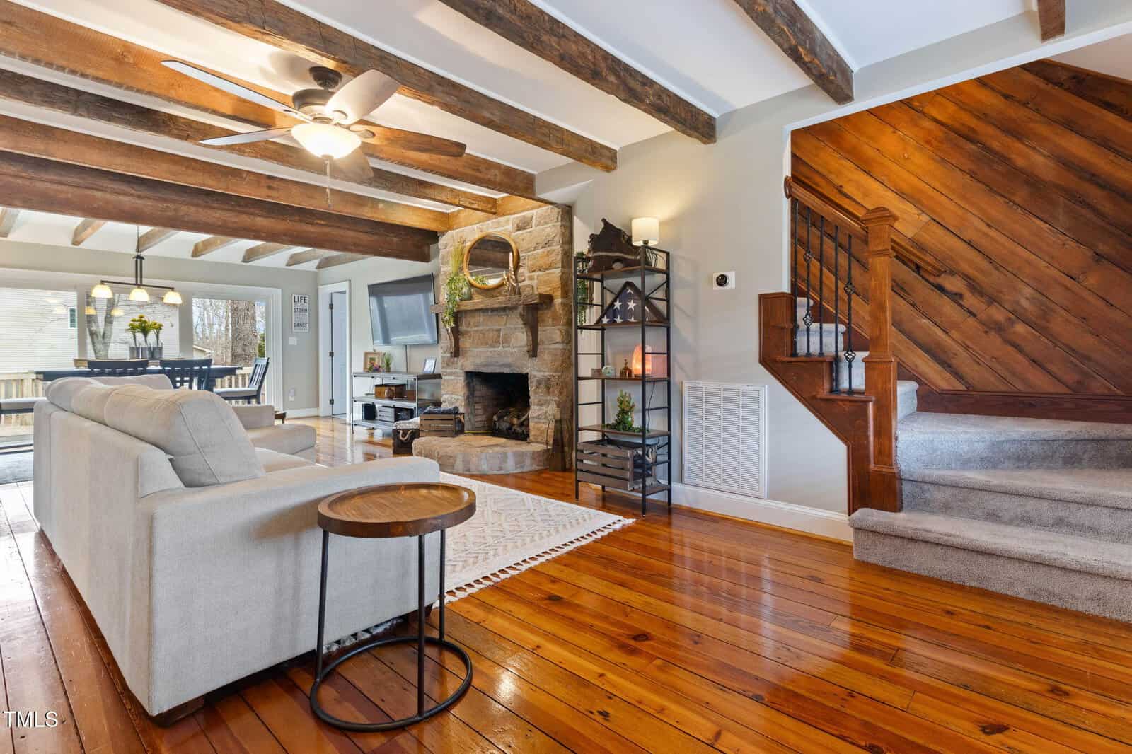 Living room with exposed wood beams, stone fireplace, wood floors, beige sectional sofa, round side table, black shelving unit with decor, and wooden staircase with carpeted steps. Dining area visible in the background.