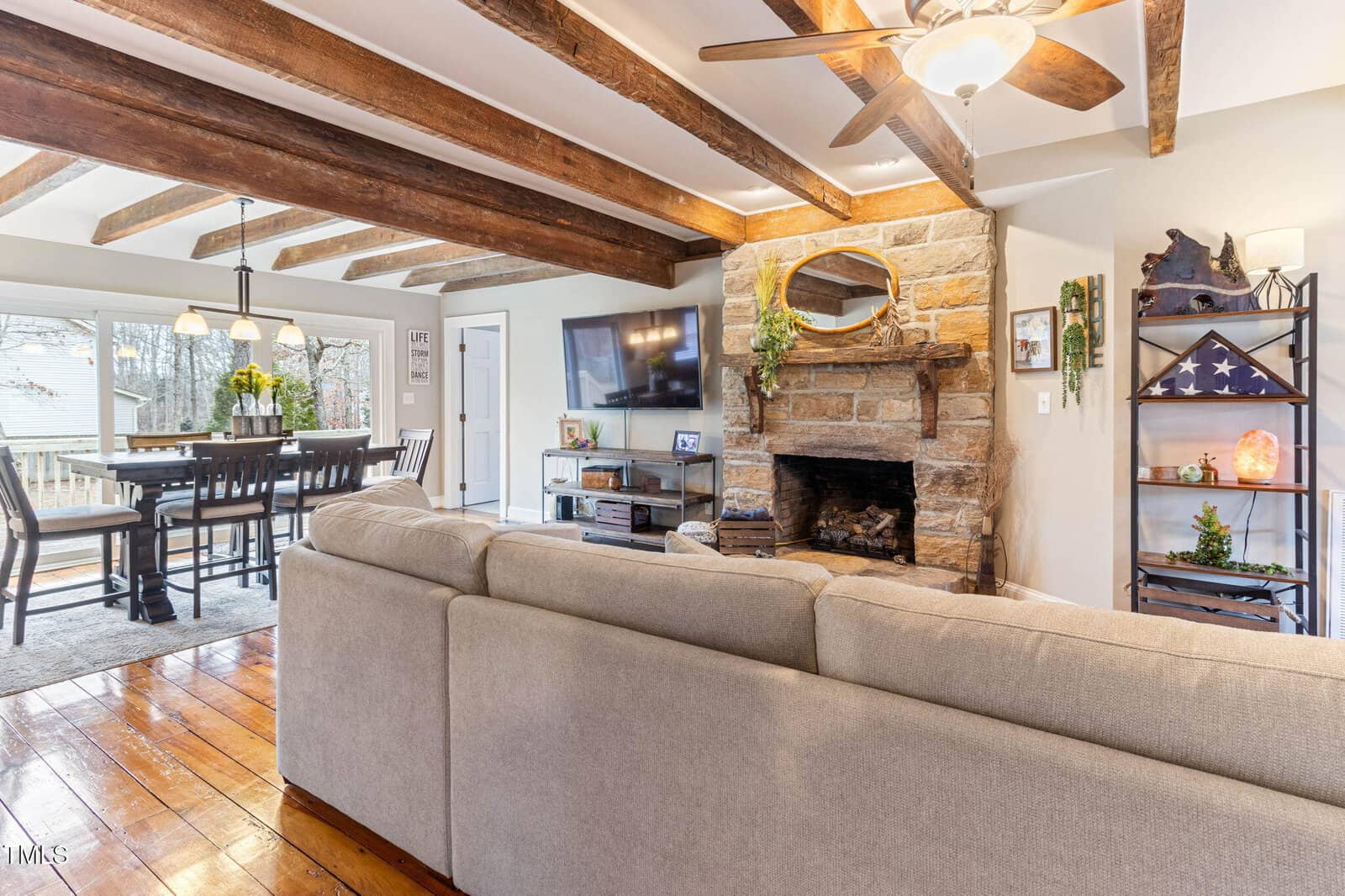 Living room with exposed wooden beams, stone fireplace, ceiling fan, and wall-mounted TV. A beige sectional faces the fireplace. Dining area with wooden table and chairs is visible in the background near large windows.