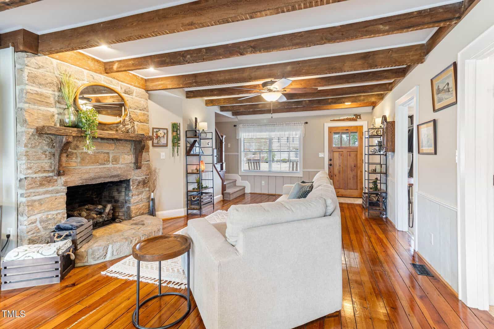 Living room with exposed wooden beams, a stone fireplace, hardwood floors, a beige sectional sofa, open shelves, a ceiling fan, and a front door with sidelights. Stairs and multiple wall art pieces are also visible.
