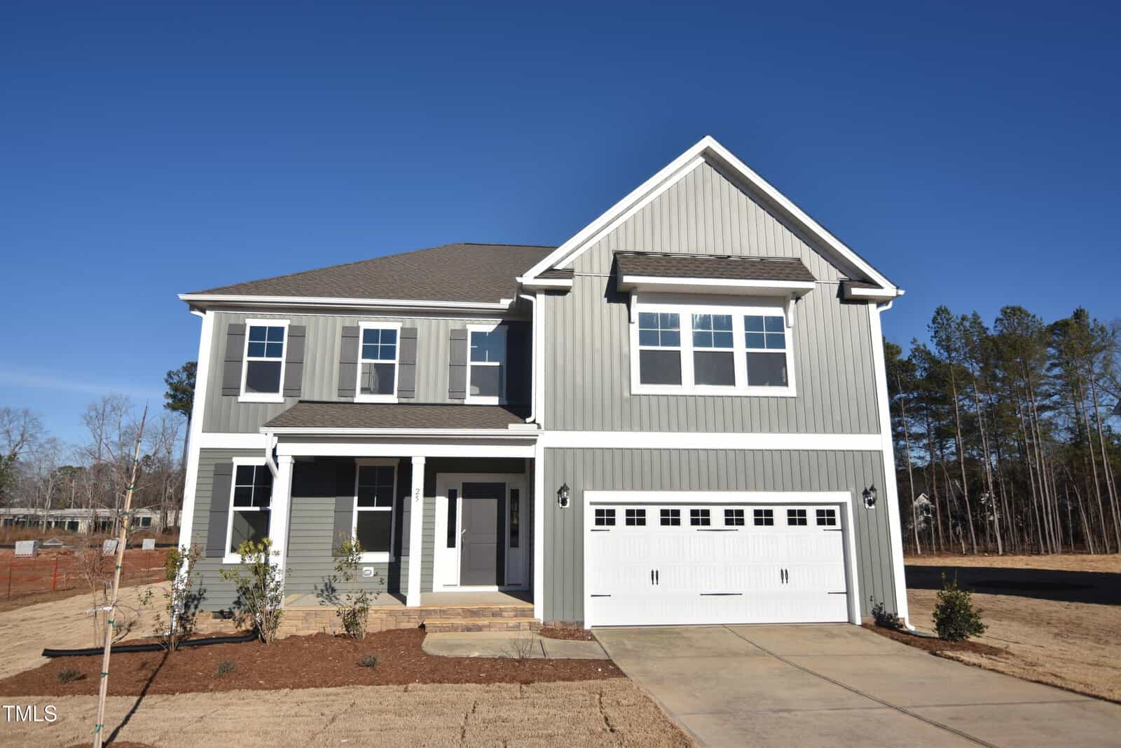 A two-story gray house with white trim features a covered front porch, double garage, and concrete driveway. The yard has minimal landscaping, with young shrubs and brown grass. Trees are visible in the background.