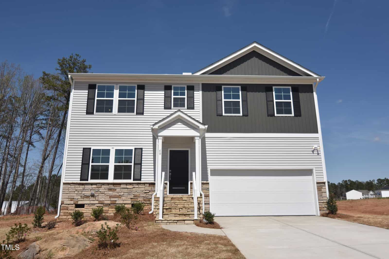 A two-story modern house with white and dark gray siding, stone accents, a covered front entrance with steps, and a white two-car garage. The yard is sparsely landscaped, and the sky is clear.