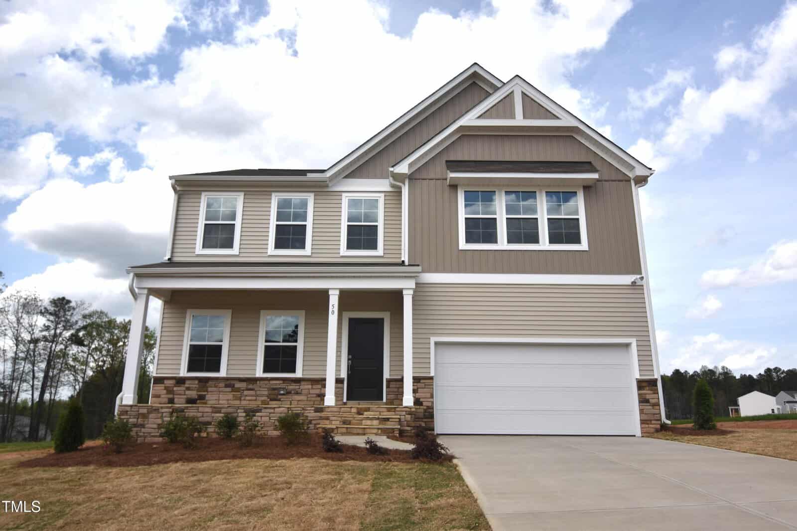 Two-story suburban house with tan siding, white trim, a covered front porch, stone accents at the base, and a double garage. The driveway and front lawn are visible, with trees and a cloudy sky in the background.
