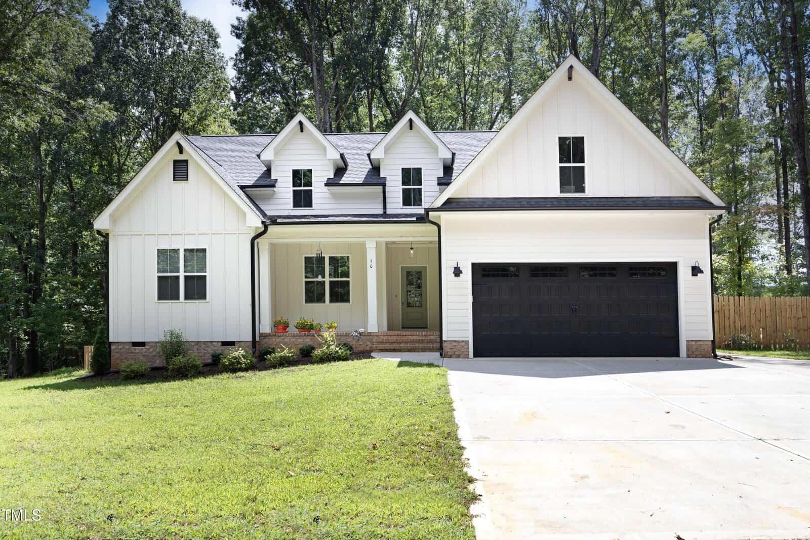 A modern white house with black trim, three dormer windows, a covered front porch with potted plants, and a two-car garage. The house is surrounded by grass lawn and bordered by tall trees.
