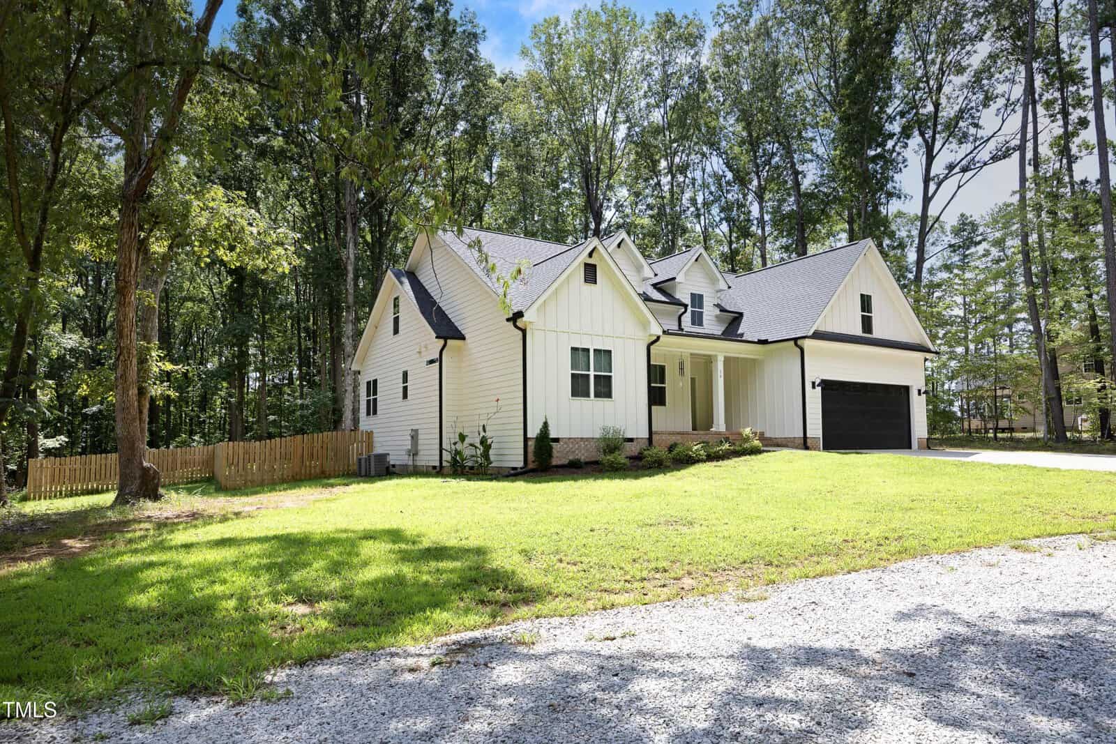 A modern white house with black trim and a dark garage door sits on a grassy lawn, surrounded by tall trees and a wooden fence. A gravel driveway leads to the front of the house. The sky is partly cloudy.