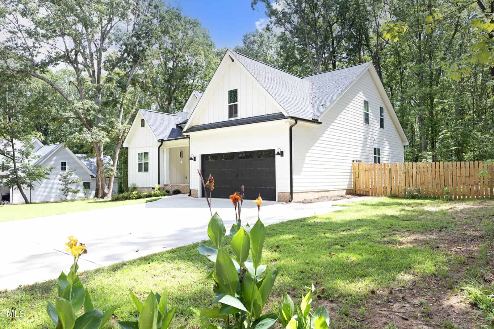 A white, modern farmhouse-style house with black accents, a double garage, and a concrete driveway. The yard has green grass, flowering plants, and a wooden fence. Trees and another house are in the background.