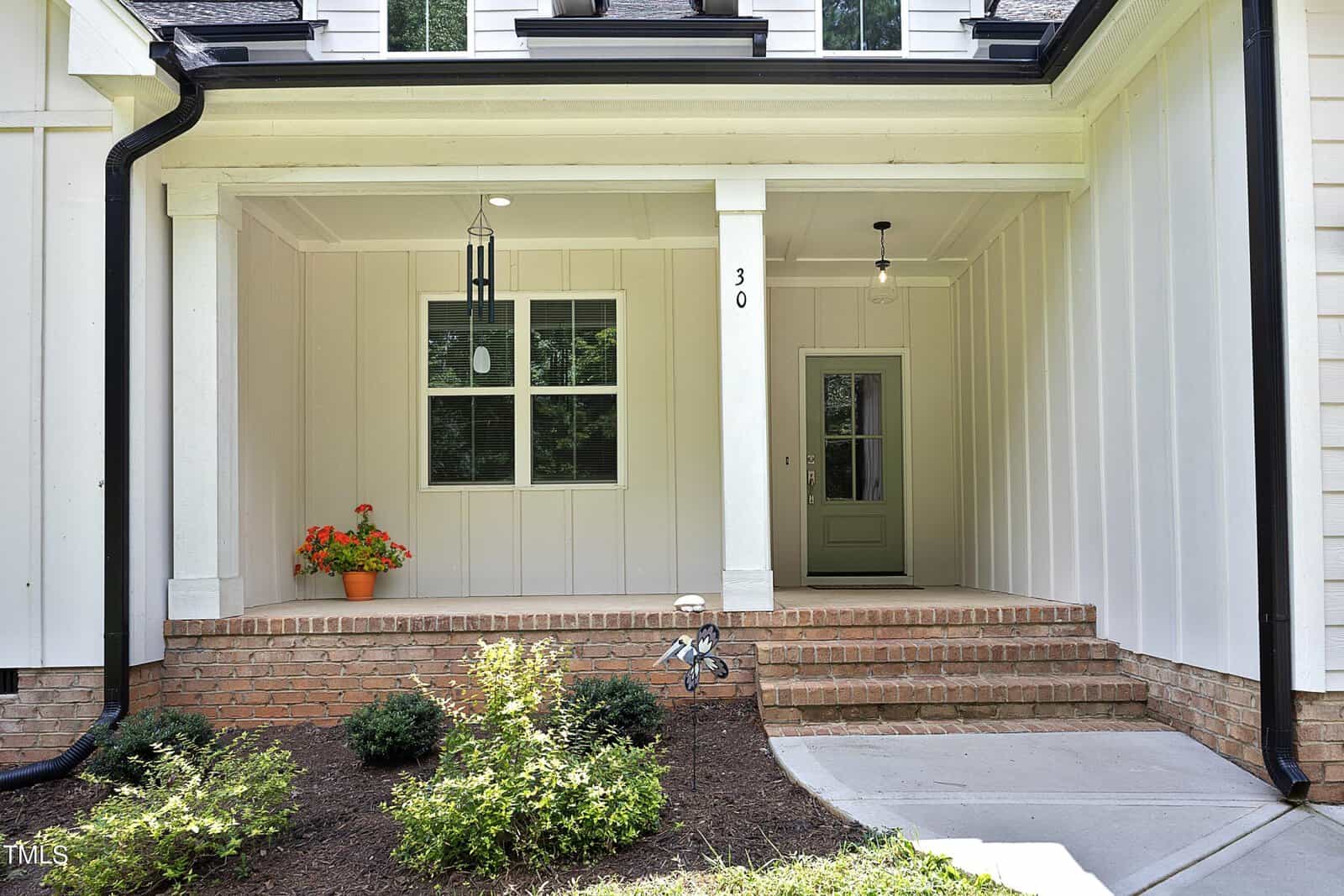 A covered front porch with white vertical siding, a single pillar, two hanging light fixtures, a green front door with glass panels, a window with an orange potted plant, brick steps, and small shrubs in the landscaping.