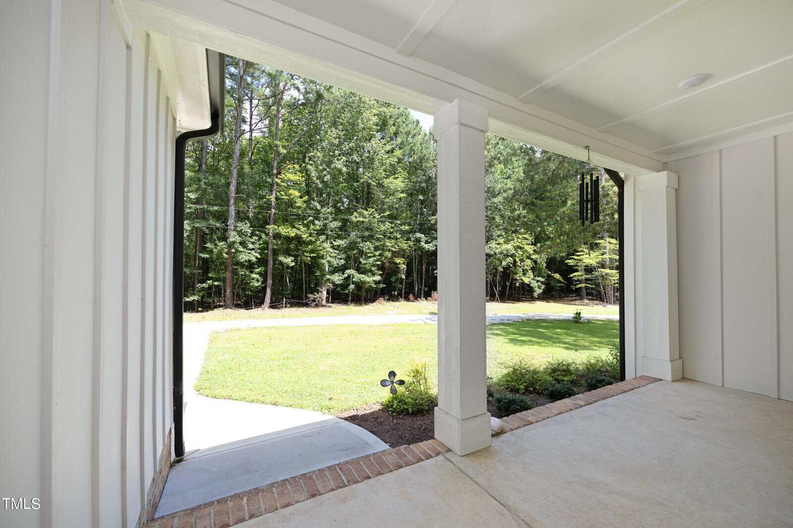 View from a covered porch with white paneling looking out onto a green lawn, a curved driveway, some landscaping, a wind chime, and a wooded area in the background.