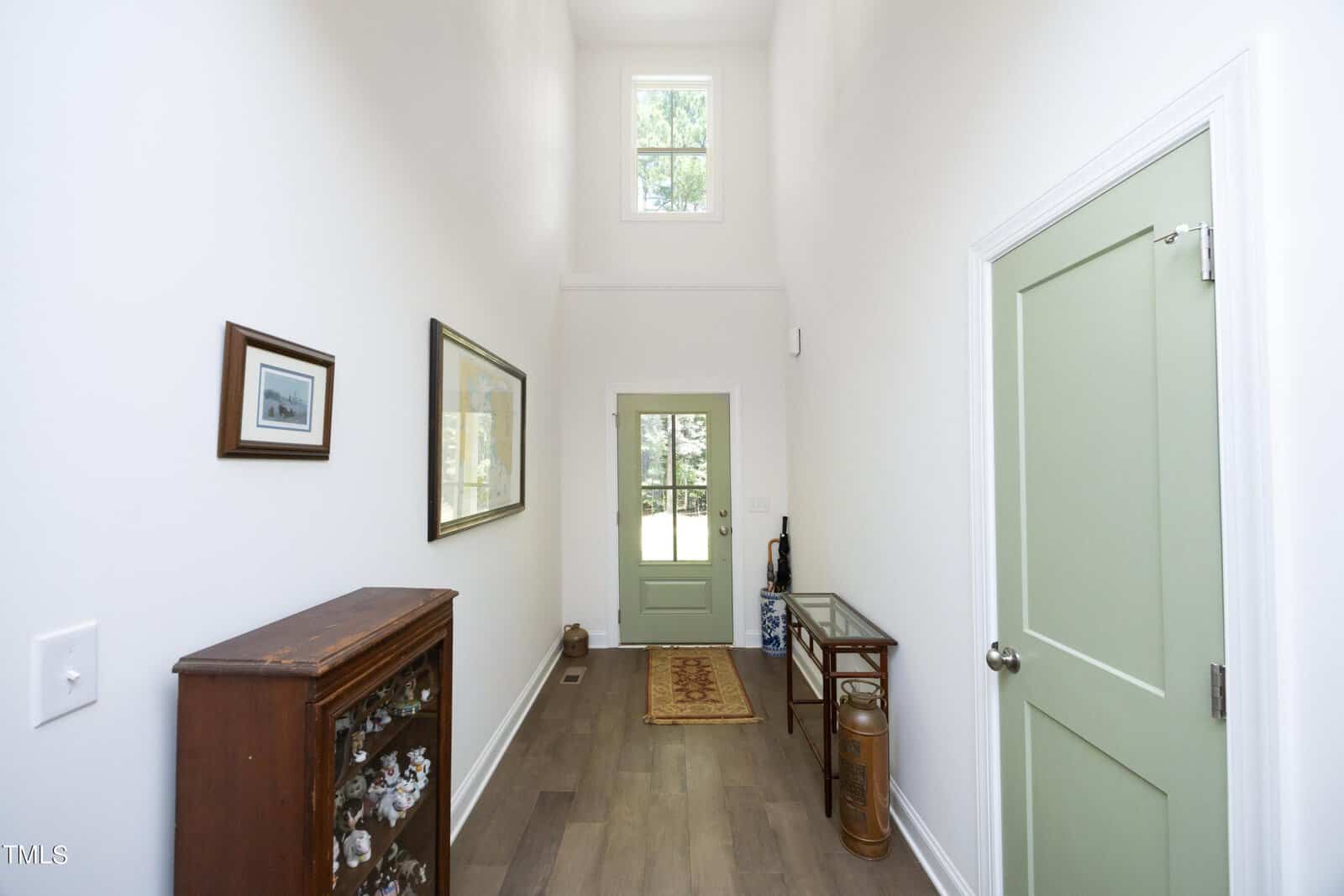 A hallway with white walls, light wood flooring, a green front door, and a high ceiling. There are framed pictures on the left wall, a small cabinet, a rug, and decorative items on a narrow glass table.