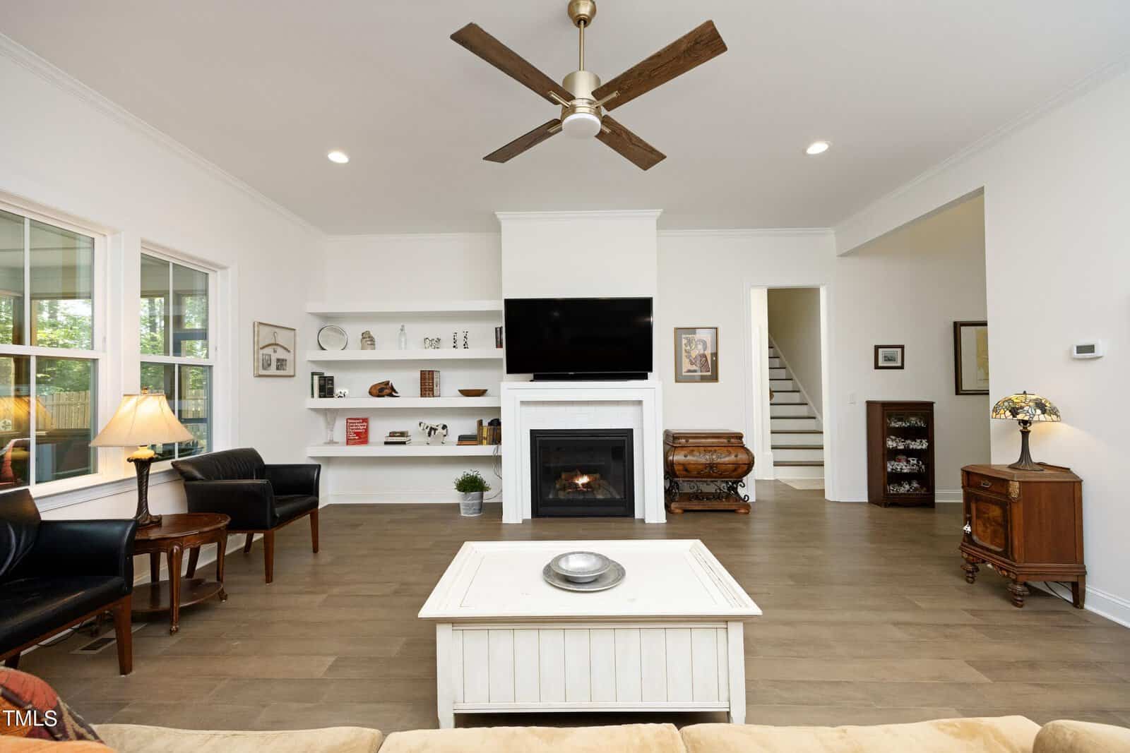 Modern living room with white walls, wood floors, a ceiling fan, fireplace with a TV above, built-in shelves, black chairs, a white coffee table, lamps, a small wooden cabinet, and staircase in the background.