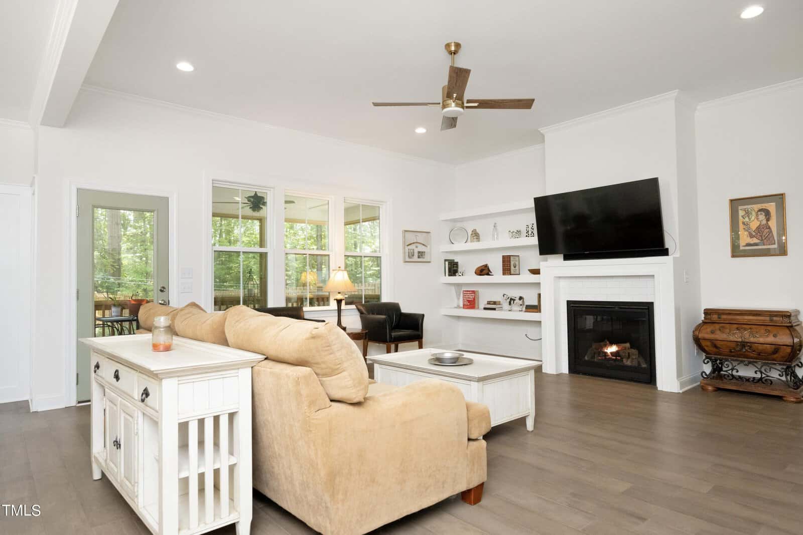 A living room with tan couches, a white coffee table, a fireplace with a TV above it, built-in shelves, and large windows overlooking a sunroom. The walls are white, and the floor is light wood.
