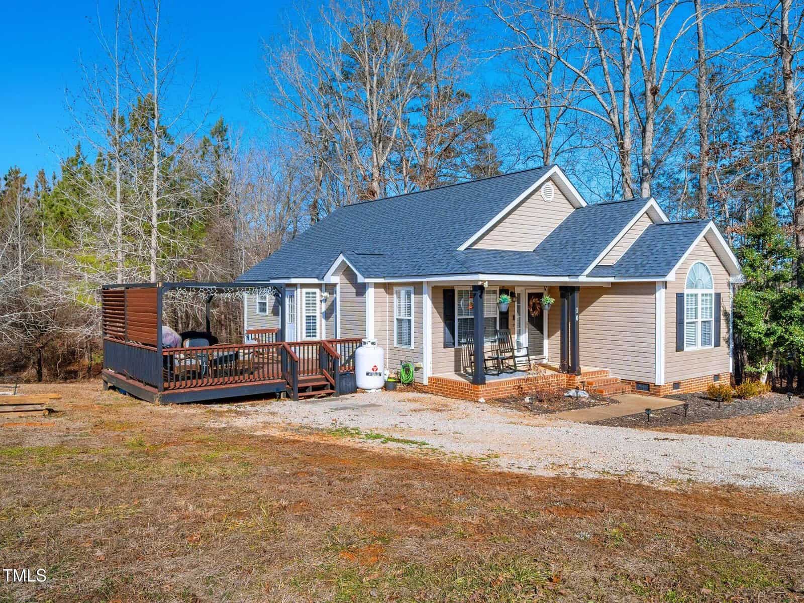 Single-story tan house with a dark roof, front porch, and attached wooden deck on the left side. The yard has sparse grass and trees in the background under a clear blue sky.