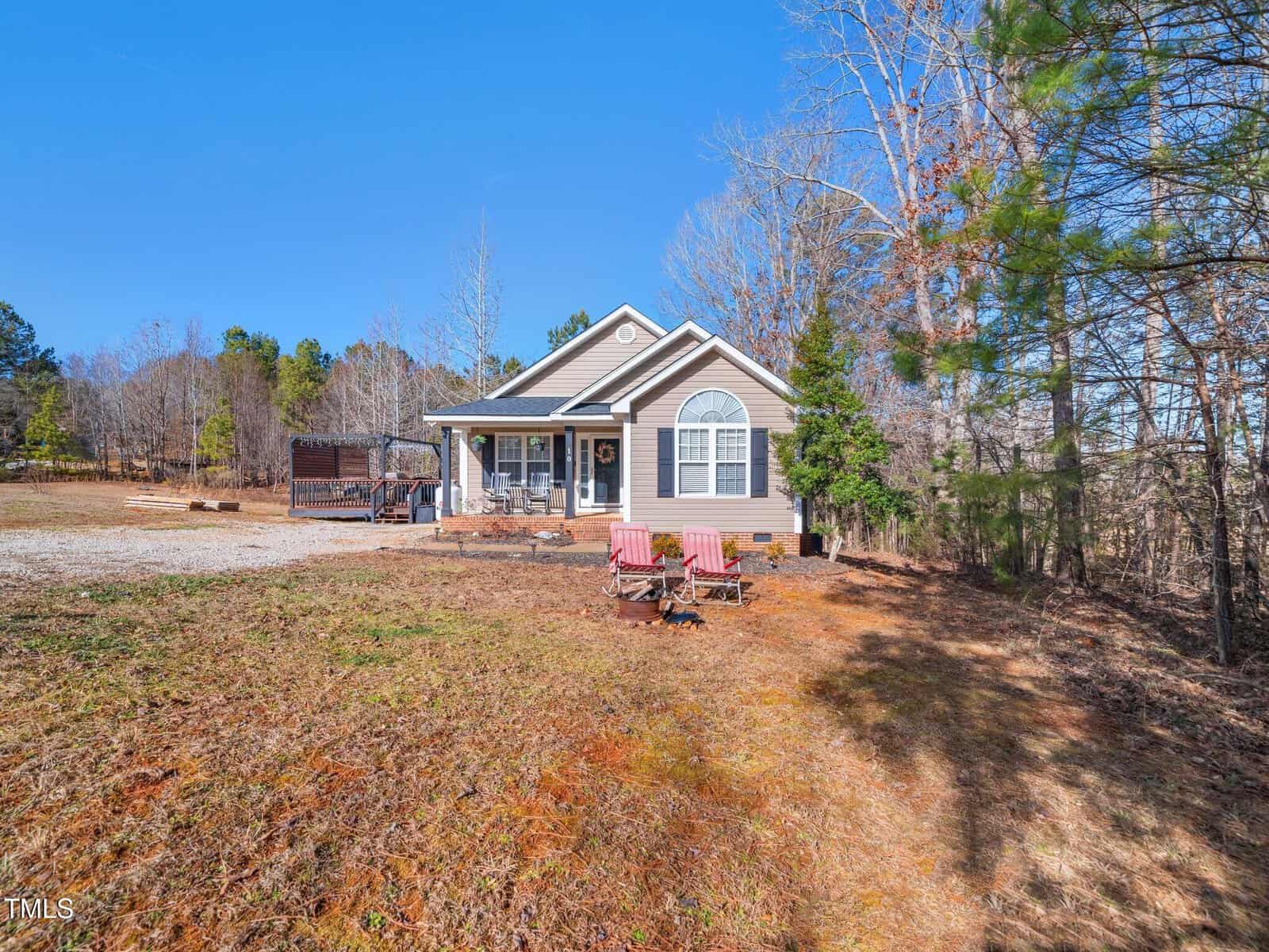 A single-story house with a covered porch sits on a large, grassy lot surrounded by trees. In front of the house are four wooden chairs arranged around a fire pit. The sky is clear and blue.