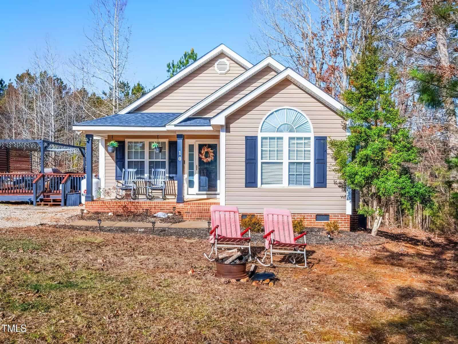 A single-story beige house with white trim and dark blue shutters features a covered porch with chairs and a wreath on the door. In the yard, two red lawn chairs face a metal fire pit. Leafless trees and some greenery surround the home.