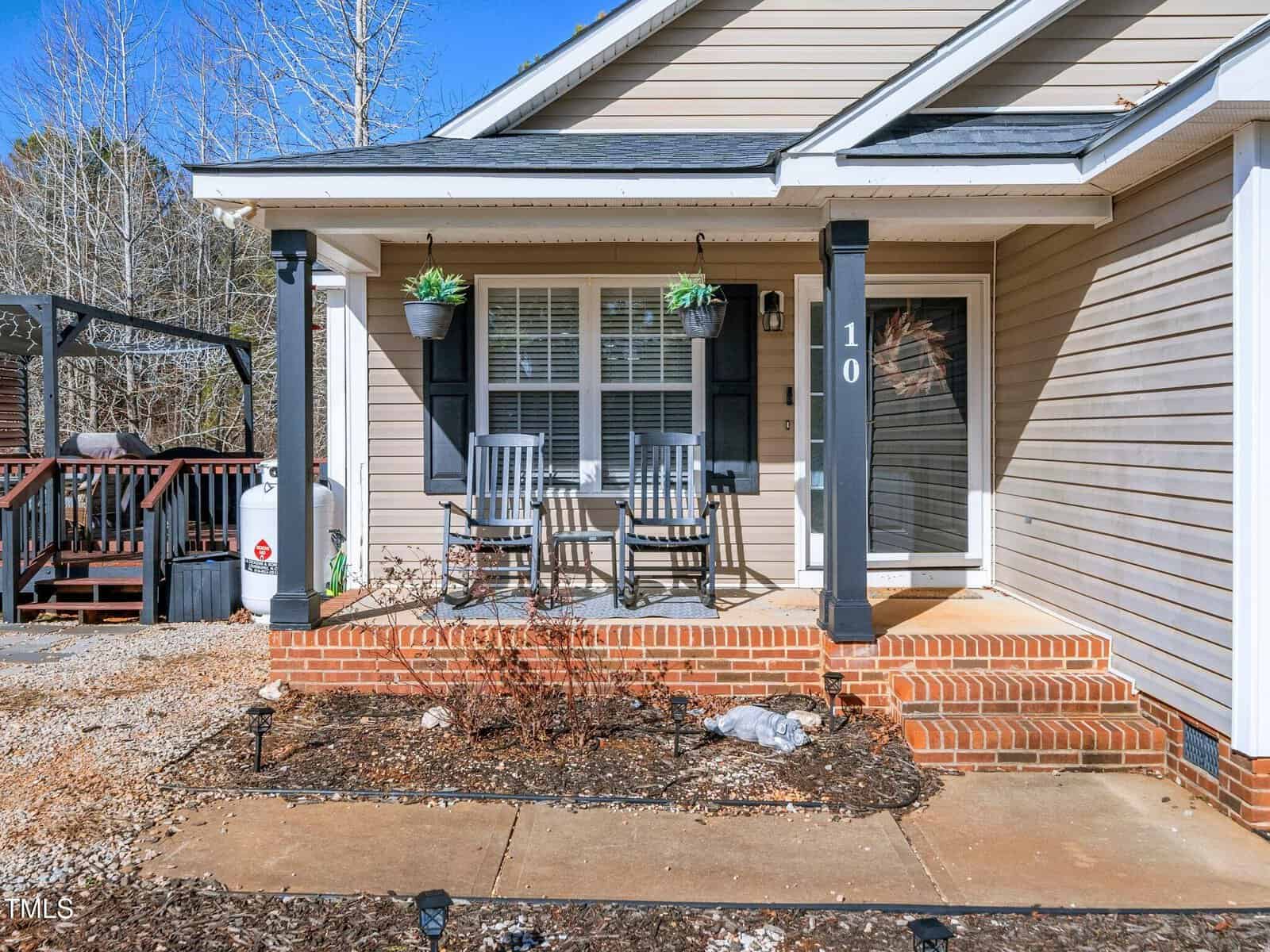 A small front porch with two black rocking chairs and a small table, hanging planters, and brick steps leading to a beige house with white trim. The area in front has mulch and bare shrubs.