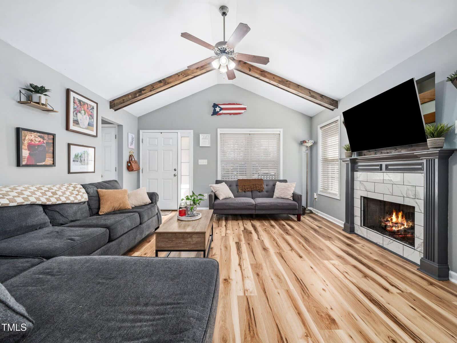 Living room with gray sofas, wood floor, fireplace with TV above, ceiling fan, exposed beam, wall art, plants, coffee table, and a Puerto Rican flag on the back wall. Bright natural light enters through windows.