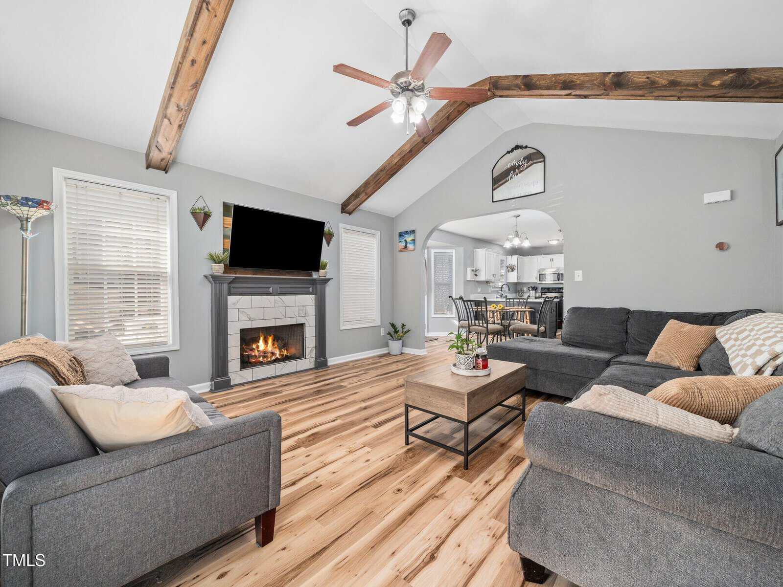 A spacious living room with wood floors, gray sofas, a coffee table, a fireplace with a TV above it, exposed wood ceiling beams, and ceiling fans, leading into a dining area and kitchen in the background.