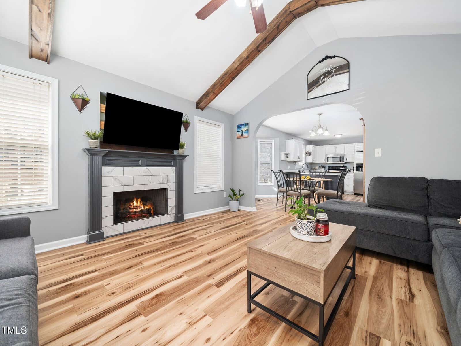 A living room with a fireplace, TV, gray sectional sofa, and wood coffee table. The space has wood floors, vaulted ceiling with exposed beams, and connects to a dining area and kitchen in the background.