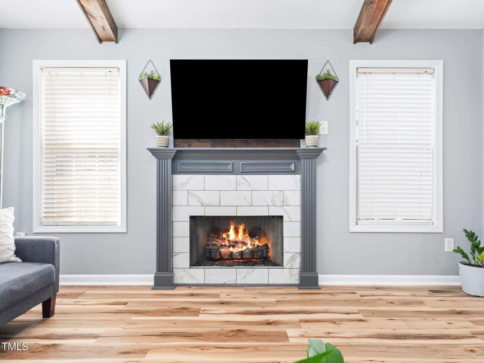 A living room features a fireplace with a fire burning, a TV mounted above, two windows with white blinds, gray walls, wood flooring, two potted plants on the mantel, and a gray couch partially visible on the left.