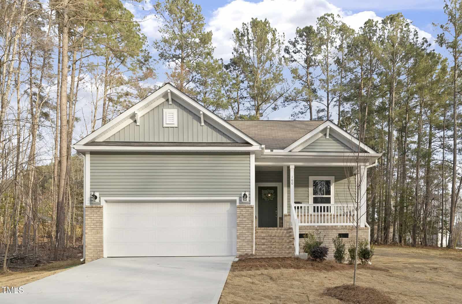 A single-story house with light green siding, a small covered front porch, white trim, and a two-car garage. The yard has sparse grass and young shrubs, with tall pine trees in the background.